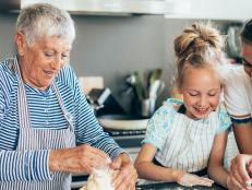 Three generation women kneading cookie dough in kitchen. Happy grandmother, mother and little girl in aprons with messy dough hands making biscuits on kitchen counter and playing with dough. Using digital tablet looking for new recipe, or watching cooking tutorial or making video vlog;