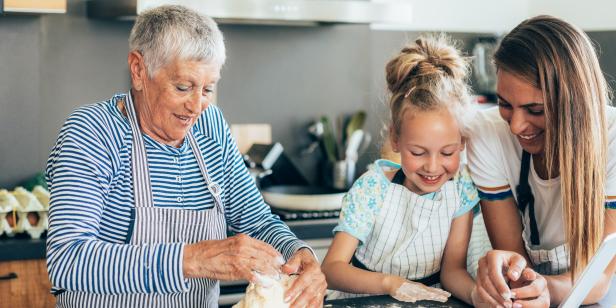 Three generation women kneading cookie dough in kitchen. Happy grandmother, mother and little girl in aprons with messy dough hands making biscuits on kitchen counter and playing with dough. Using digital tablet looking for new recipe, or watching cooking tutorial or making video vlog;