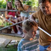 A shot of a young son with his father, the father has made a homemade pizza and he's placing the pizza on a cutting board using a pizza peel, the young son is excited to eat and the mother and daughter are watching in the distance.