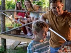 A shot of a young son with his father, the father has made a homemade pizza and he's placing the pizza on a cutting board using a pizza peel, the young son is excited to eat and the mother and daughter are watching in the distance.