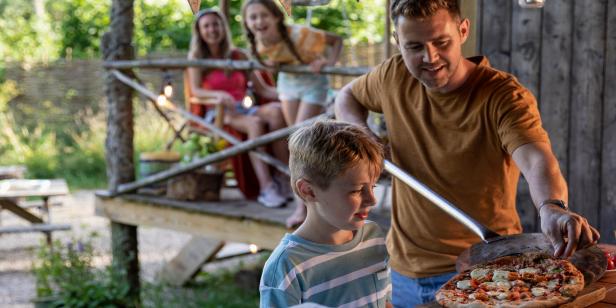 A shot of a young son with his father, the father has made a homemade pizza and he's placing the pizza on a cutting board using a pizza peel, the young son is excited to eat and the mother and daughter are watching in the distance.