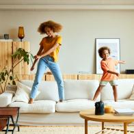 Full length of happy mother and son dancing on sofa in living room. Woman and boy enjoying at home. They are in casuals.