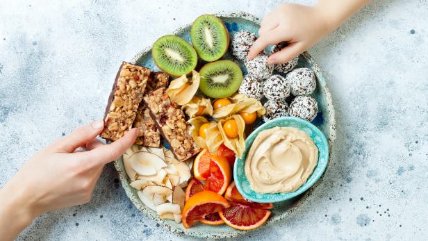 Mother sharing healthy vegan dessert snacks with toddler child. Concept of healthy sweets for children. Protein granola bars, homemade raw energy balls, cashew butter, toasted coconut chips, fruits platter
