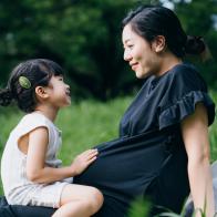 Lovely little Asian girl gently touching her pregnant mother's baby. Big sister talking to the baby and feels the movement of baby in the belly of mother while sitting on meadow in the nature. Sibling love. Expecting a new life with love and care concept