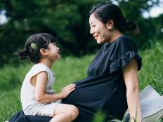 Lovely little Asian girl gently touching her pregnant mother's baby. Big sister talking to the baby and feels the movement of baby in the belly of mother while sitting on meadow in the nature. Sibling love. Expecting a new life with love and care concept