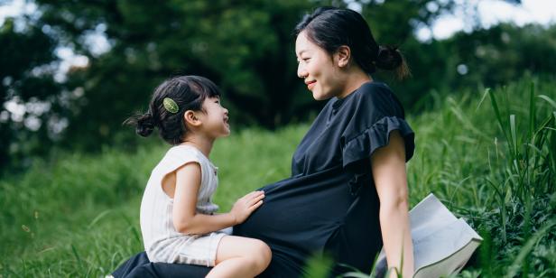 Lovely little Asian girl gently touching her pregnant mother's baby. Big sister talking to the baby and feels the movement of baby in the belly of mother while sitting on meadow in the nature. Sibling love. Expecting a new life with love and care concept