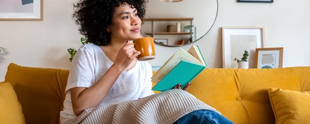 Pensive relaxed African american woman reading a book at home, drinking coffee sitting on the couch. Copy space. Lifestyle concept.