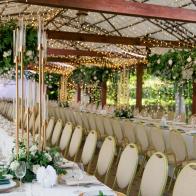 Stylish wooden terrace is prepared for the luxurious wedding. Tables are decorated with greenery and high golden candlesticks. Whole area is illuminated with yellow electric garlands.