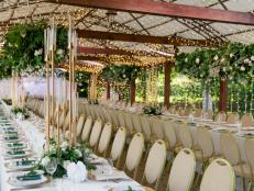 Stylish wooden terrace is prepared for the luxurious wedding. Tables are decorated with greenery and high golden candlesticks. Whole area is illuminated with yellow electric garlands.
