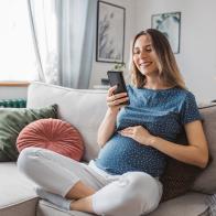 Young pregnant woman at home. She is sitting on sofa and using smart phone for online shopping or to pay bills.