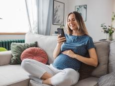 Young pregnant woman at home. She is sitting on sofa and using smart phone for online shopping or to pay bills.