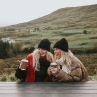 Two young woman holding takeaway coffee cups look cozy, wrapped up warmly in puffer jackets, hats and scarves, and snuggled up close to eachother. They smile and chat. One points at something.