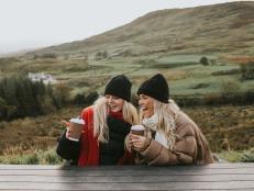 Two young woman holding takeaway coffee cups look cozy, wrapped up warmly in puffer jackets, hats and scarves, and snuggled up close to eachother. They smile and chat. One points at something.