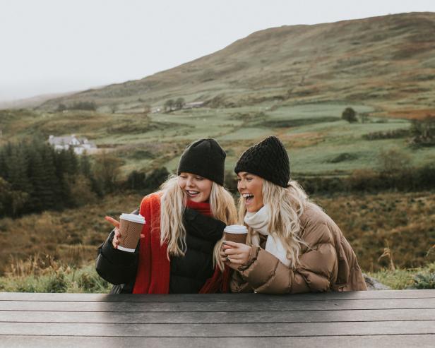 Two young woman holding takeaway coffee cups look cozy, wrapped up warmly in puffer jackets, hats and scarves, and snuggled up close to eachother. They smile and chat. One points at something.