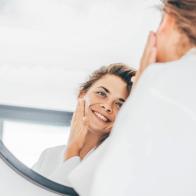 Young woman with long curly hair in bathrobe enjoys daily routine applying moisturizing cream on face in bedroom at home closeup mirror reflection