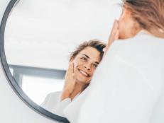 Young woman with long curly hair in bathrobe enjoys daily routine applying moisturizing cream on face in bedroom at home closeup mirror reflection