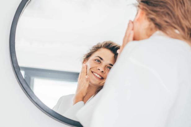 Young woman with long curly hair in bathrobe enjoys daily routine applying moisturizing cream on face in bedroom at home closeup mirror reflection