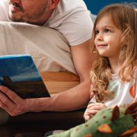 Portrait of a little girl and her father in the morning. He is lying on the bad face down while she is sitting on the floor. He is smiling while holding a book and reading a morning story to her.