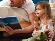 Portrait of a little girl and her father in the morning. He is lying on the bad face down while she is sitting on the floor. He is smiling while holding a book and reading a morning story to her.