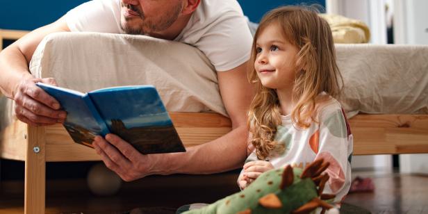 Portrait of a little girl and her father in the morning. He is lying on the bad face down while she is sitting on the floor. He is smiling while holding a book and reading a morning story to her.