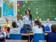 An Ethnic teacher is leading a class of elementary school children. There are various posters on the wall, and drawings on the chalkboard. Students are putting up their hands to answer a question.