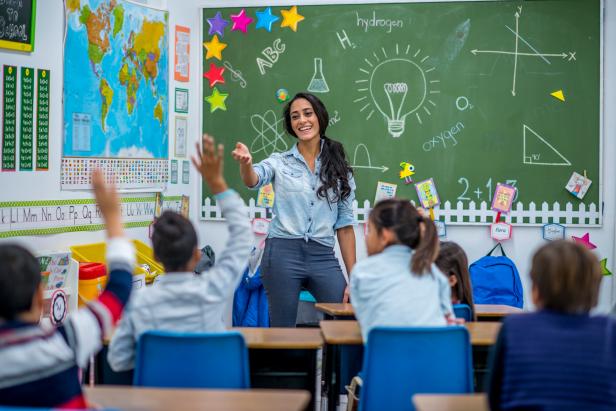 An Ethnic teacher is leading a class of elementary school children. There are various posters on the wall, and drawings on the chalkboard. Students are putting up their hands to answer a question.