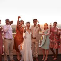 Portrait of happy just married couple and guests toasting drinks while standing against sky at sunset