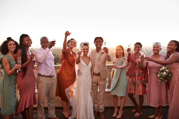 Portrait of happy just married couple and guests toasting drinks while standing against sky at sunset