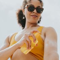 Waist up image of a woman wearing sunglasses, spraying suncream on to her arm.