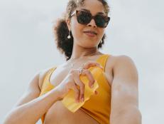 Waist up image of a woman wearing sunglasses, spraying suncream on to her arm.