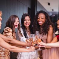 A happy group of diverse adult female friends toasting to their Latin friend who is about to get married soon. They are all cheering and looking excited. The girls are dressed in fancy dresses and are located in a luxurious apartment.