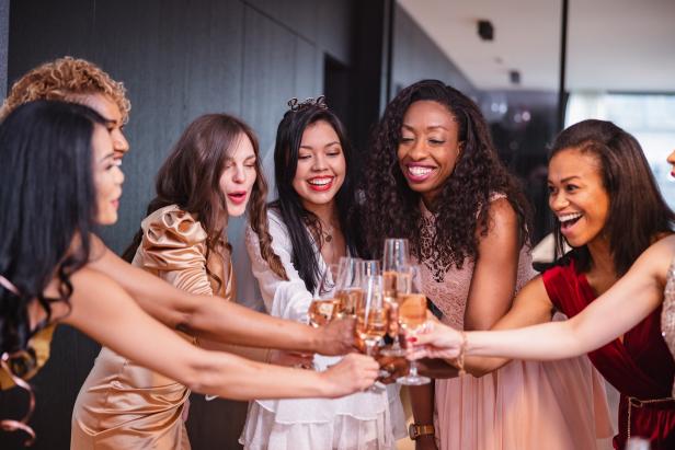 A happy group of diverse adult female friends toasting to their Latin friend who is about to get married soon. They are all cheering and looking excited. The girls are dressed in fancy dresses and are located in a luxurious apartment.