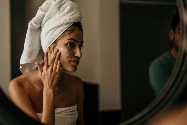 A young woman applies moisturizer to her face, her reflection in the mirror capturing her dedication to self-care