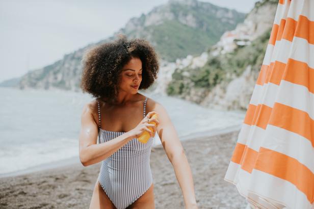 A woman on a beach uses a spray bottle to spritz liquid sun cream / sunscreen over her body.