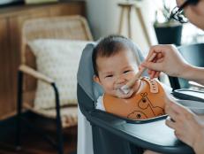Asian mother feeding messy baby girl with spoon in high chair at home, cheeky baby girl smiling and looking at camera. Baby milestone, growth and development