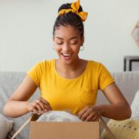 Quick Delivery Service Concept. Happy black lady received package, unpacking cardboard box, sitting on the sofa in living room at home, copy space. Female buyer satisfied with online shopping purchase