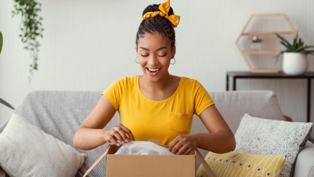 Quick Delivery Service Concept. Happy black lady received package, unpacking cardboard box, sitting on the sofa in living room at home, copy space. Female buyer satisfied with online shopping purchase