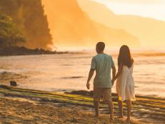 A middle-aged Eurasian couple stroll along a beautiful Hawaii beach at sunset. The waves are rolling in on the right side and the forested mountains are rising up ahead of them as they walk away from the camera.