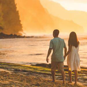 A middle-aged Eurasian couple stroll along a beautiful Hawaii beach at sunset. The waves are rolling in on the right side and the forested mountains are rising up ahead of them as they walk away from the camera.