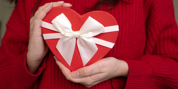 Red heart-shaped gift box with white ribbon in woman's hands, close-up, concept of Valentine's Day present.