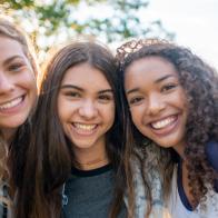 A small group of three female friends huddle in closely together as they pose for a portrait.  They are each dressed casually and are smiling on the warm summers evening.