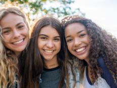 A small group of three female friends huddle in closely together as they pose for a portrait.  They are each dressed casually and are smiling on the warm summers evening.