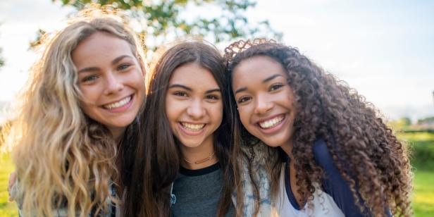 A small group of three female friends huddle in closely together as they pose for a portrait.  They are each dressed casually and are smiling on the warm summers evening.