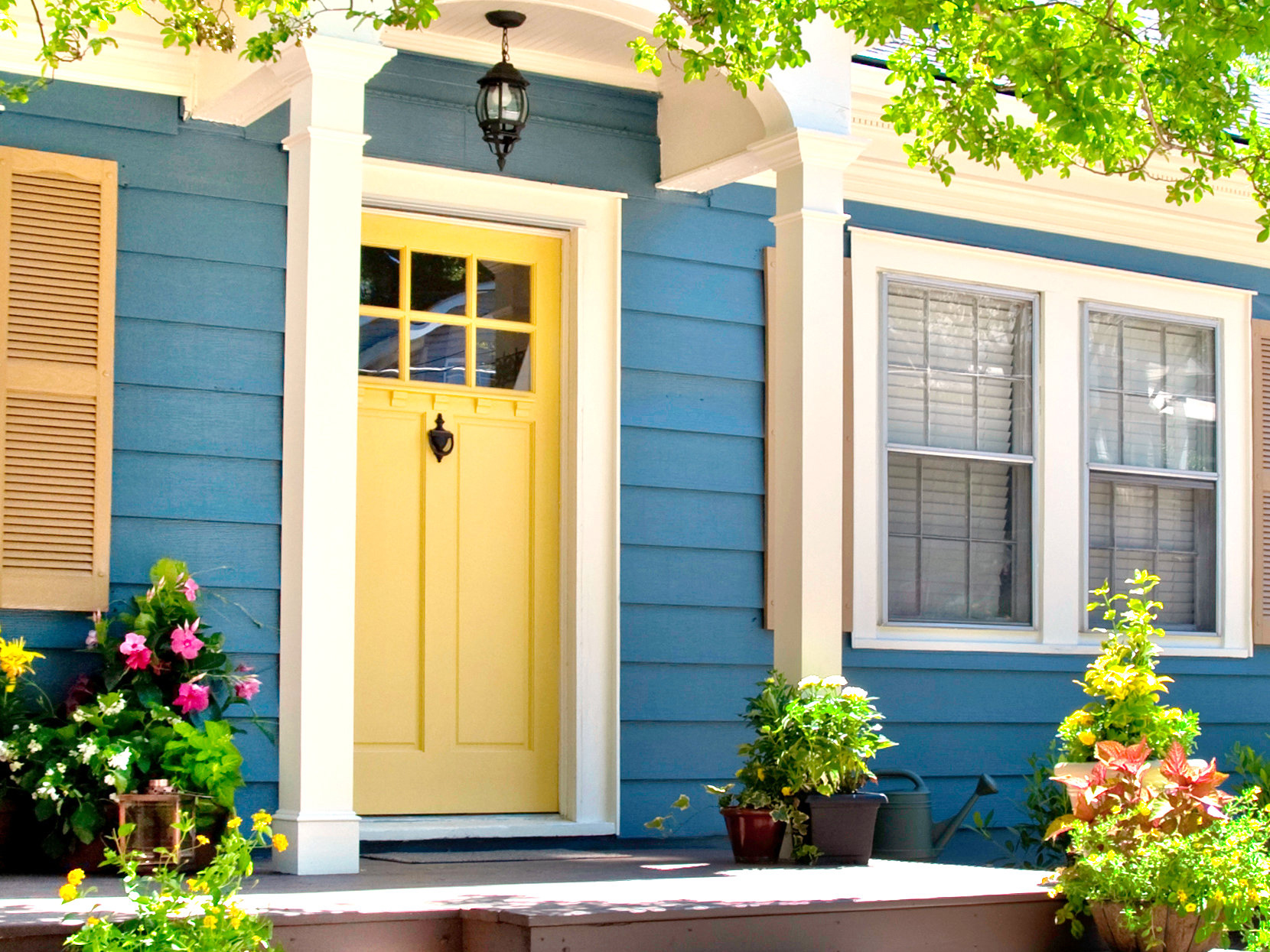 Blue house exterior with yellow door, new windows, flowers, plants, and covered porch area. 